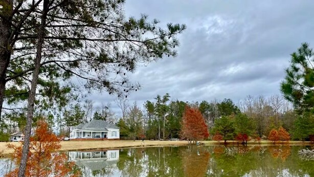 Una casa junto a un lago tranquilo, rodeada de árboles y con follaje otoñal bajo un cielo nublado.