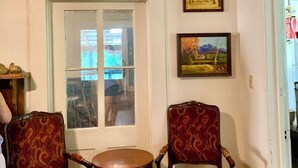Una habitación con dos sillones ornamentados y una pequeña mesa redonda, una ventana con persianas, y dos cuadros enmarcados en la pared.