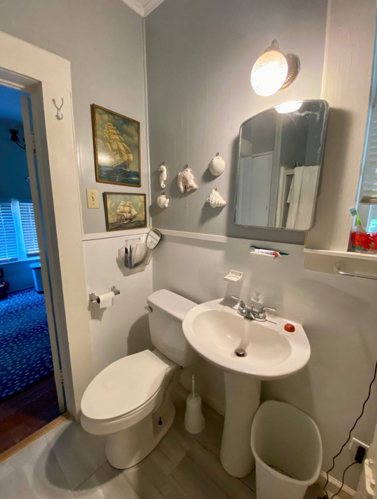 Un baño con inodoro, lavamanos y espejo. Hay conchas decorativas en la pared y un cuadro sobre el inodoro.