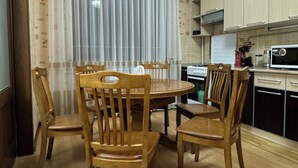 Un coin repas avec une table et des chaises en bois, un lustre, et une cuisine avec des placards et des appareils électroménagers en arrière-plan.