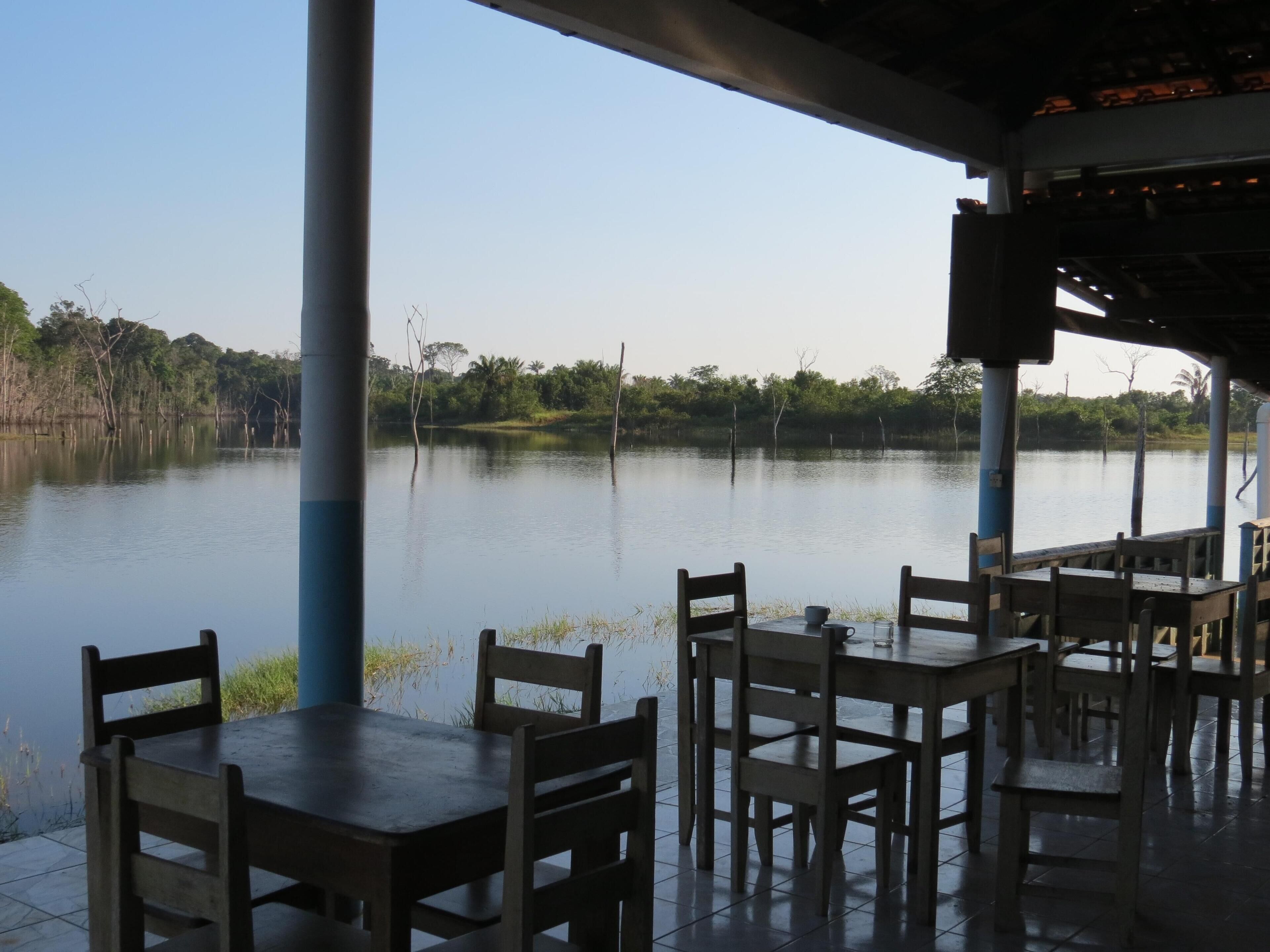 Restaurante com área externa e mesas de frente para um lago tranquilo, cercado por árvores.