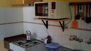 A kitchen with a gas stove, a kettle, a microwave on a shelf, and various utensils hanging on the wall. - Condor de los Andes (El Chalten)
