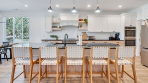 A modern kitchen with white cabinets, a central island with bar stools, pendant lighting, and a view of the outdoors through large windows.