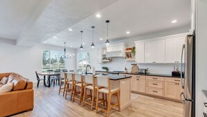 A modern kitchen with white cabinets, a dining area with wooden chairs, and a large window providing natural light.