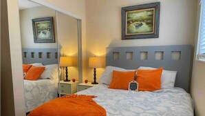 Ein Doppelbett mit einem gemusterten Überwurf und orangefarbenen Kissen, ein Nachttisch mit Lampe und ein Spiegel, der den Raum widerspiegelt.