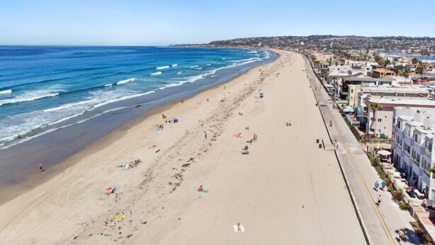 Ein Strand mit einer weiten Sandfläche, vereinzelten Sonnenschirmen und Gebäuden entlang der Küste.