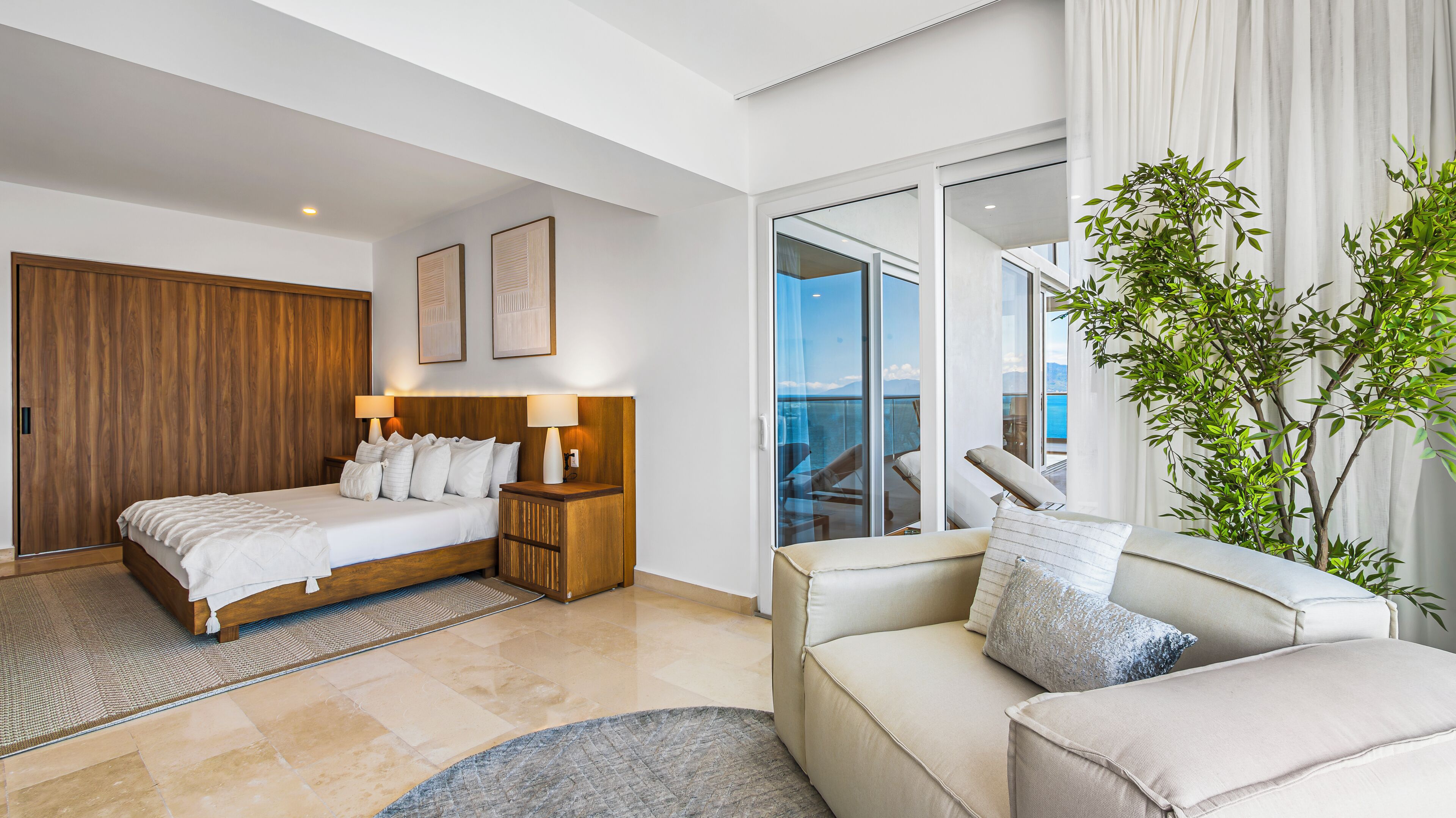 A modern hotel room with a bed, a sofa, a balcony with a view, and a large plant.