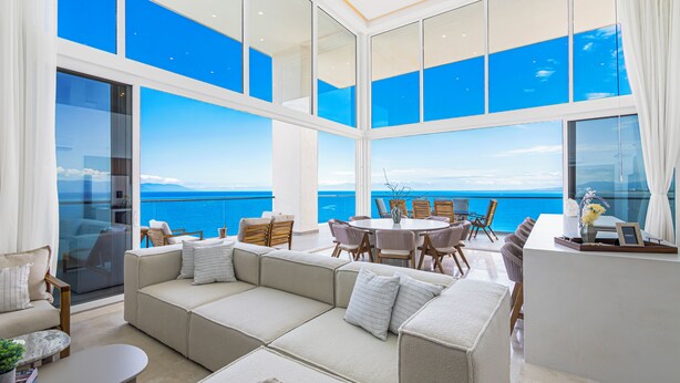 A modern interior with a large sofa, dining table, and chairs, overlooking a scenic ocean view.