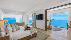 A spacious bedroom with a bed, a TV, a balcony with a view of the sea, and a dining area.