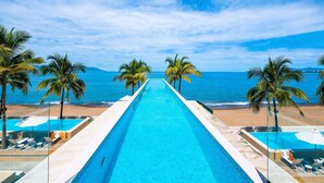 A long swimming pool with clear blue water, surrounded by palm trees and a beach view.