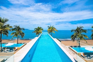 Una piscina larga con agua cristalina azul, rodeada de palmeras y vistas a la playa.