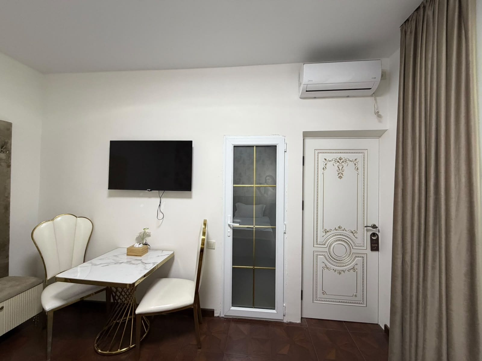 A room with a white table, two chairs, a television mounted on the wall, a sliding glass door, and an air conditioning unit.