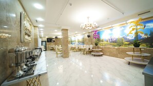 Eine Hotelhalle mit Marmorboden, einem Buffetbereich mit Essensausgabegeräten und einem Wandgemälde, das eine Berglandschaft darstellt.