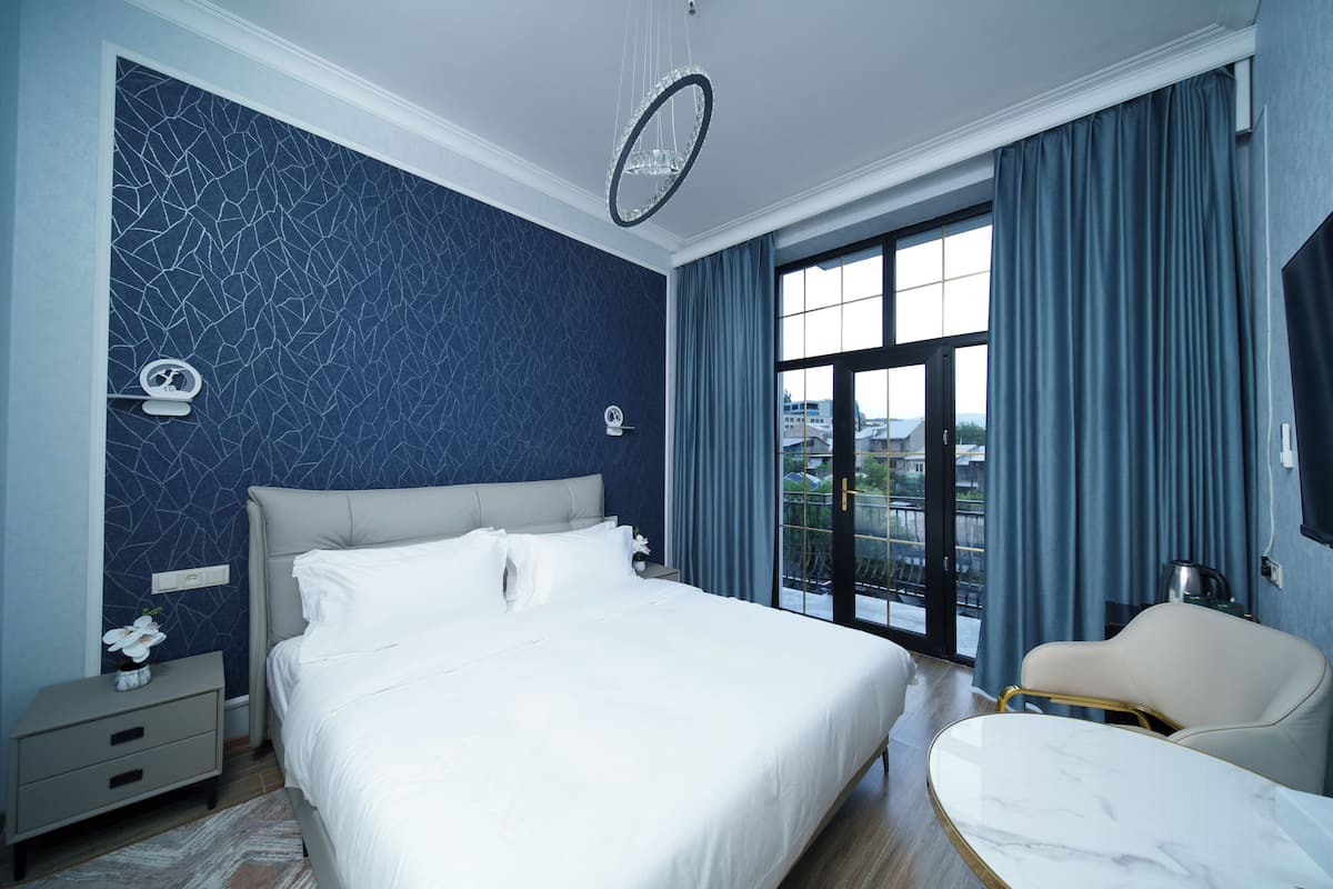 A modern bedroom with a large bed, a nightstand, a chair, and a view of the outdoors through a large window.