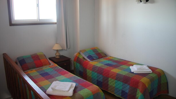 A bedroom with two single beds, each with a colorful quilt, a nightstand with a lamp, and a window with curtains. - Gemelos Apart (Villa Gesell)