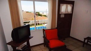 A living room with a television on a wooden stand, two red chairs, a small table, and a view of a swimming pool through a window. - Gemelos Apart (Villa Gesell)