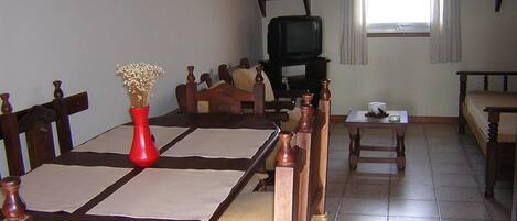 Un comedor con una mesa de madera, sillas y un jarrón rojo con flores. Se ve un televisor al fondo y una ventana con cortinas.