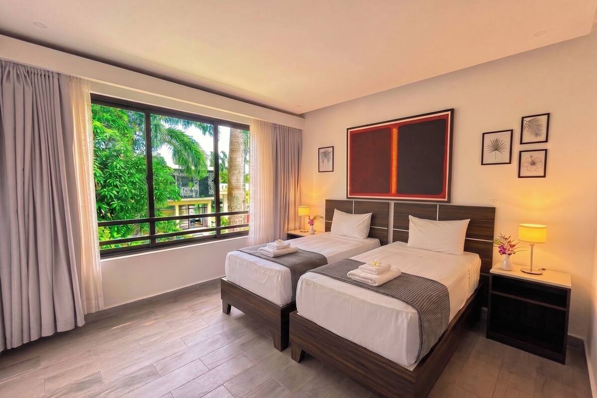 A hotel room with two beds, a large window with curtains, a bedside table with a lamp, and framed artwork on the wall.