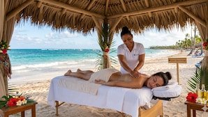 A beach massage setup with a massage table, a masseuse, and a sign indicating a beach massage service. - Hotel Neon (Punta Cana)