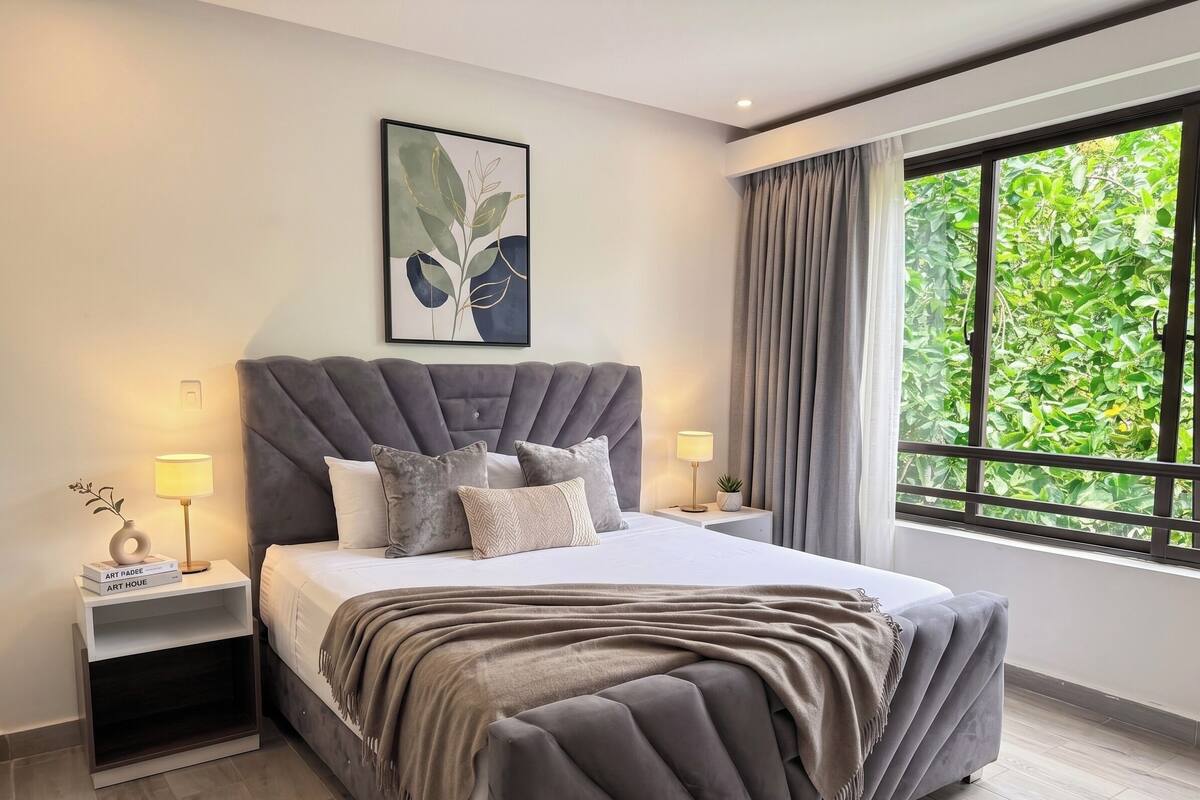 A modern bedroom with a tufted headboard, a large window with a view of greenery, a bedside table with a lamp, and a bedside plant.