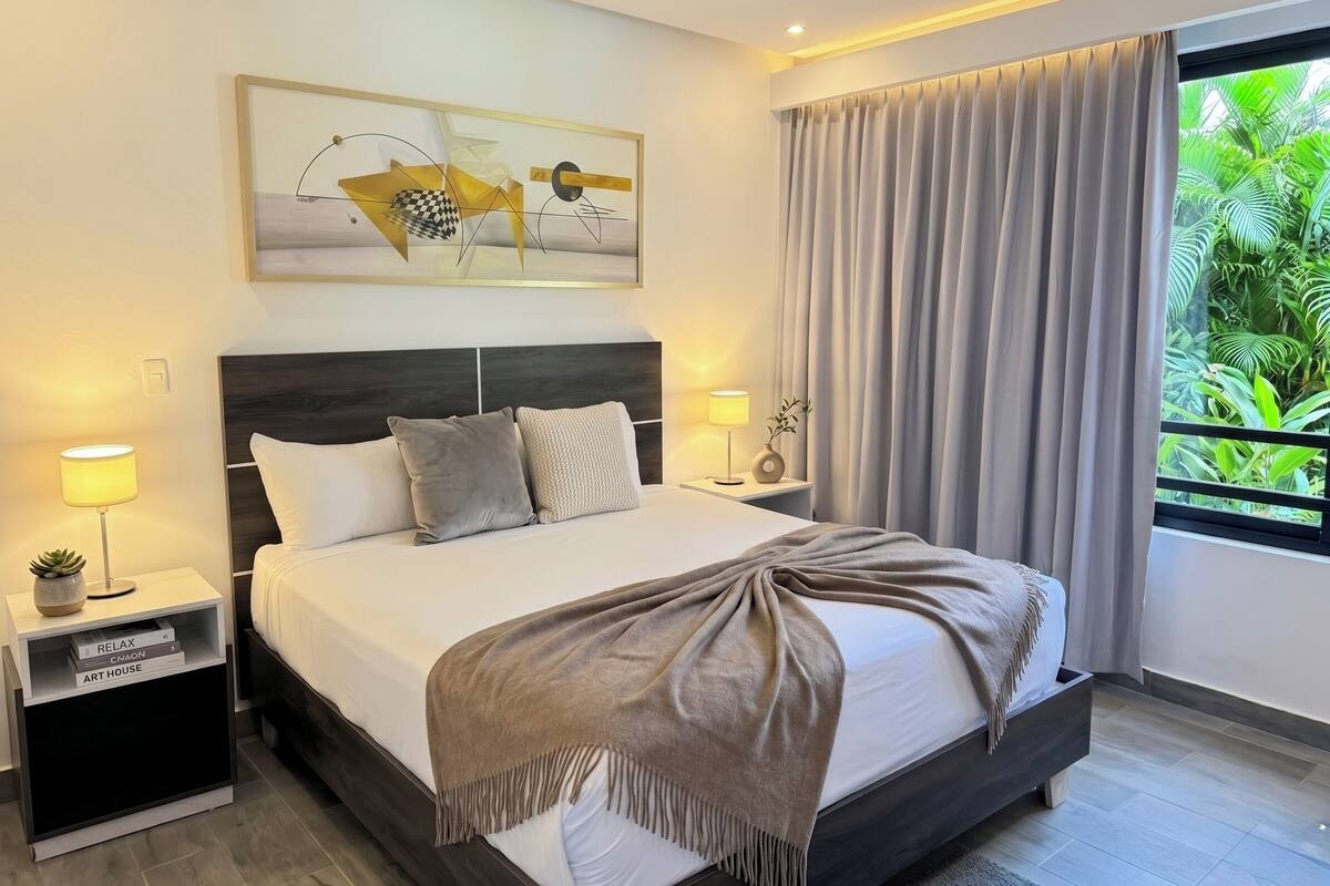 A modern hotel room with a large bed, two bedside tables, a nightstand, a framed abstract painting, a window with curtains, and a view of greenery outside.