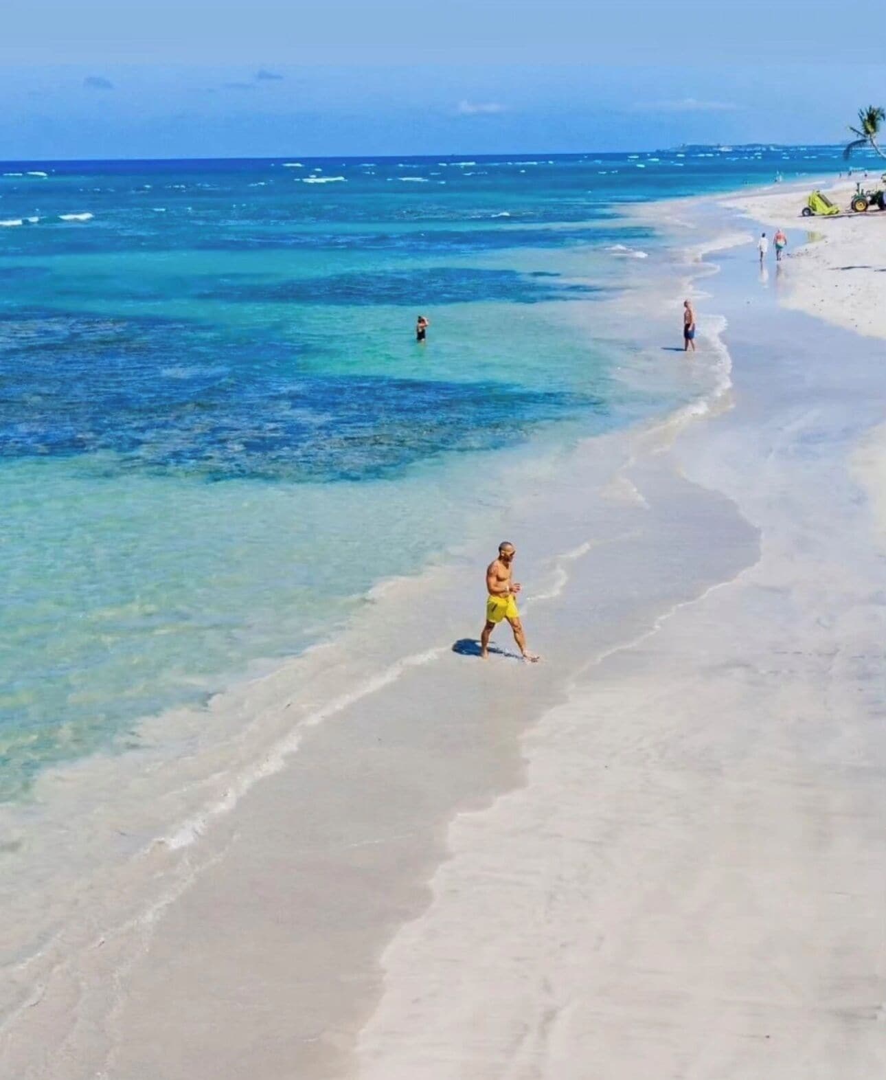 A beach with clear turquoise water, a sandy shore, and a few people walking.