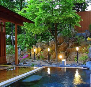 Un área de piscina tranquila al aire libre con una terraza de madera, una formación rocosa y un árbol grande que da sombra.