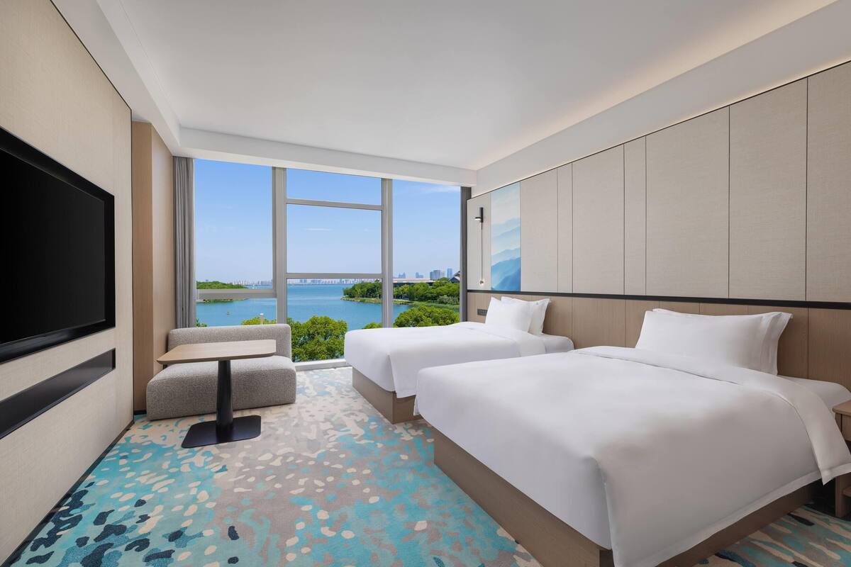 A hotel room with two beds, a TV, a small table, and a view of a body of water and cityscape.