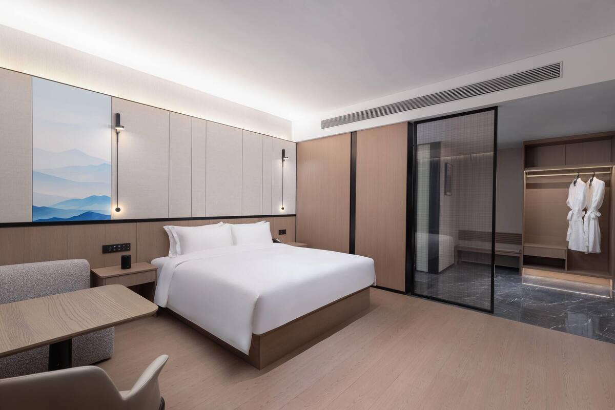 A modern hotel room with a large bed, a bedside table, a seating area, and a wardrobe.