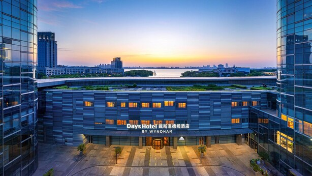 A modern hotel with a flat roof garden, large glass windows, and a view of the city skyline at sunset. - Days Hotel By Wyndham Suzhou Dushu Lake (Suzhou)