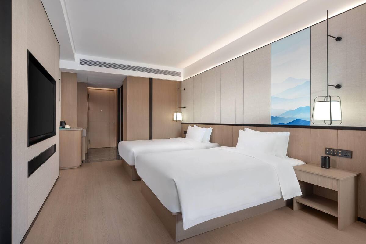 A hotel room with two beds, a TV, a bedside table, and a large window with a mountain view.