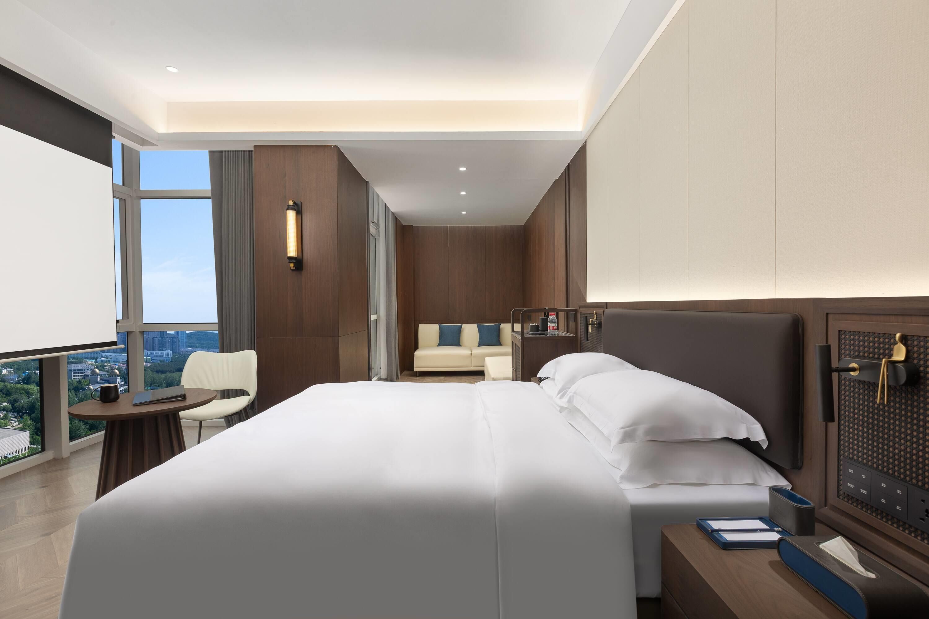 Une chambre d’hôtel moderne dotée d’un grand lit, d’un bureau avec une chaise, d’une petite table et offrant une vue sur la ville grâce à de grandes fenêtres.