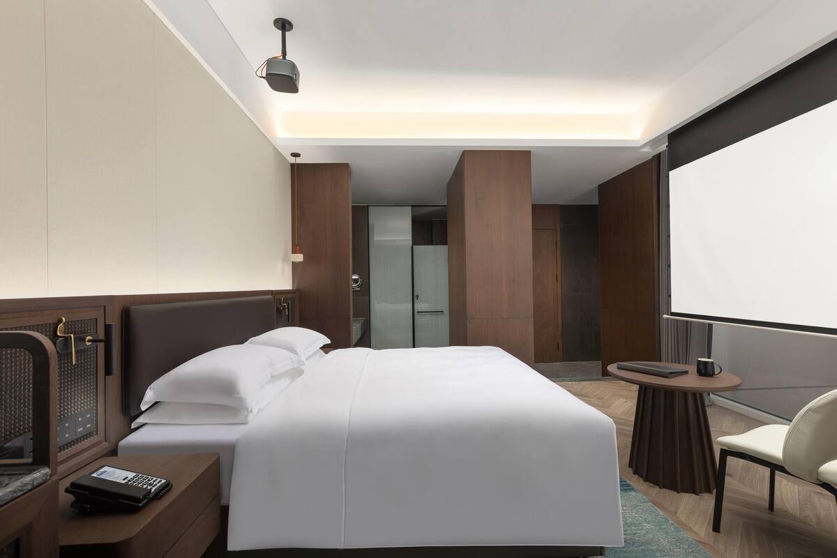 A modern hotel room with a large bed, a desk with a laptop, a chair, and a large window.