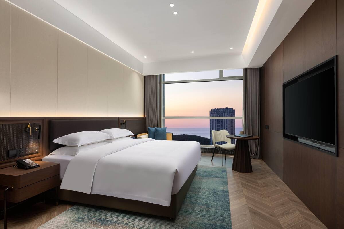 A modern hotel room with a large bed, a desk with a phone, a TV, and a view of a cityscape through large windows.