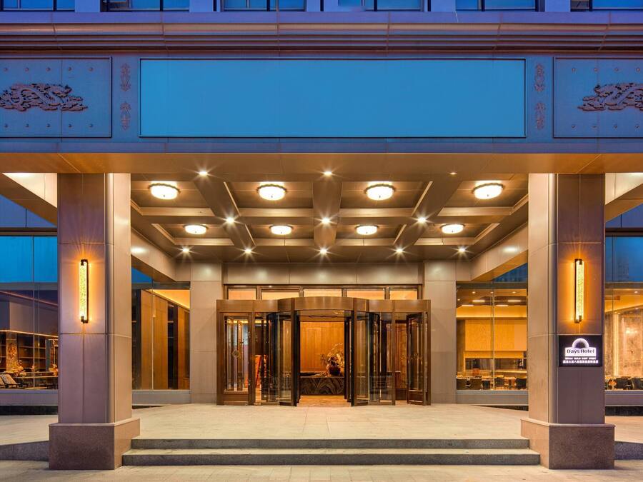 Una entrada de hotel con diseño moderno, que presenta un acceso por puerta de vidrio, un área de espera techada y un letrero con el nombre del hotel.