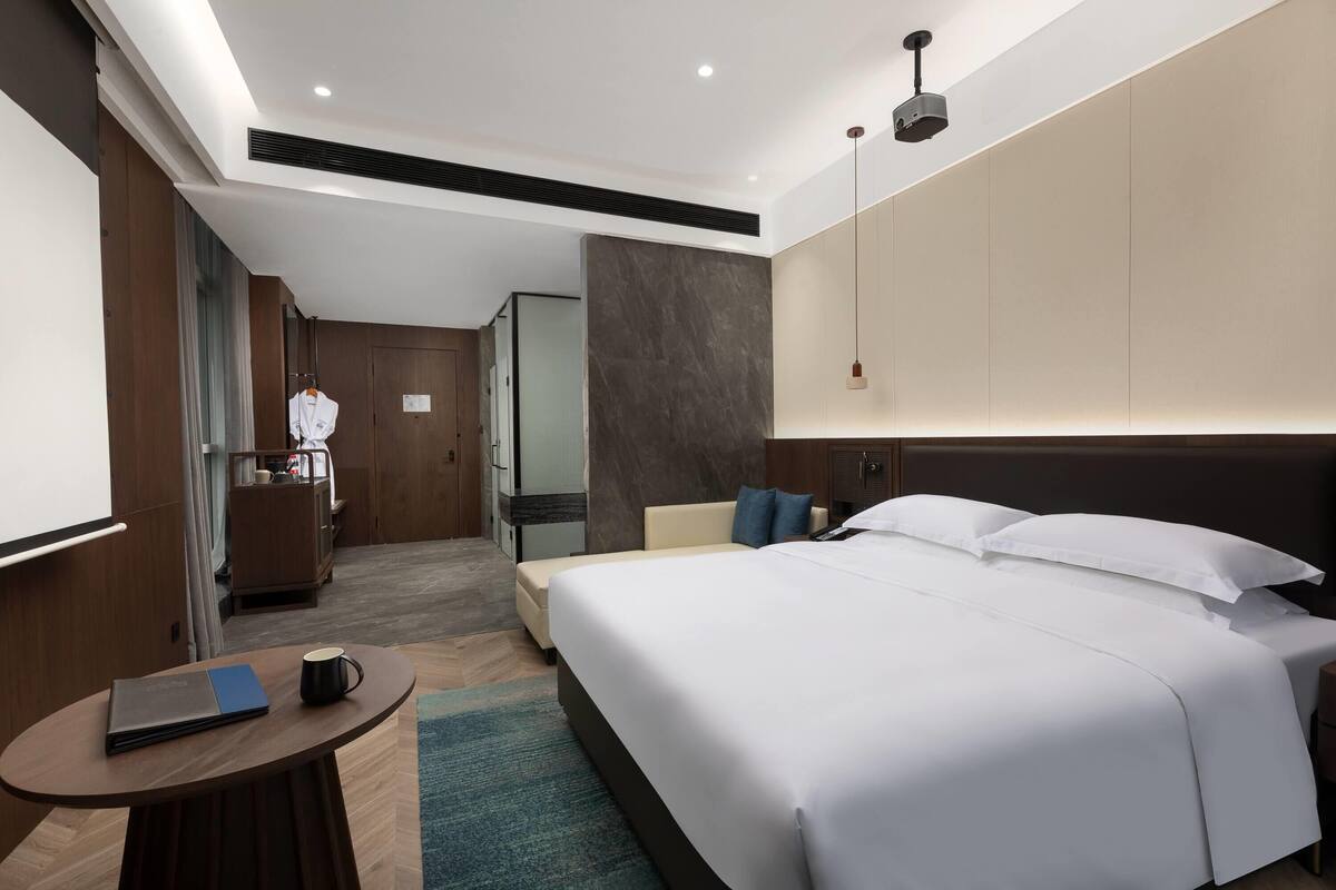 A hotel room with a large bed, a bedside table with a book and a mug, a small round table, and a view of the hallway.