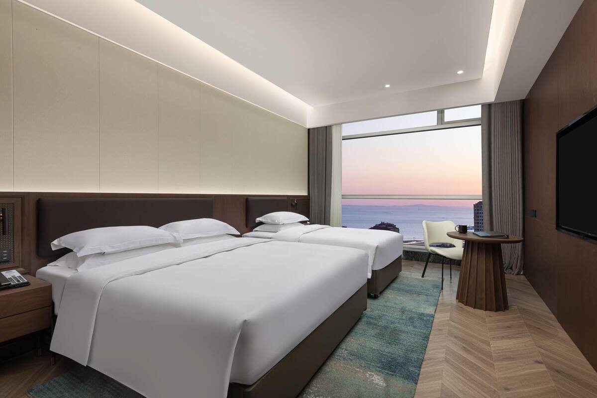 A hotel room with two beds, a desk, a chair, a TV, and a view of the ocean.