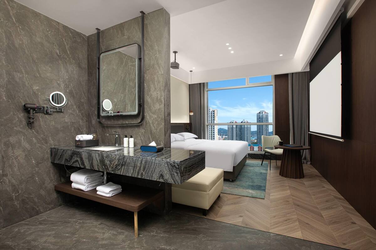 A modern hotel room with a marble bathroom, a large bed, a seating area, and a city view.