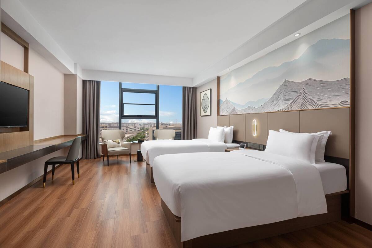 A hotel room with two beds, a desk, a chair, and a large window offering a city view.