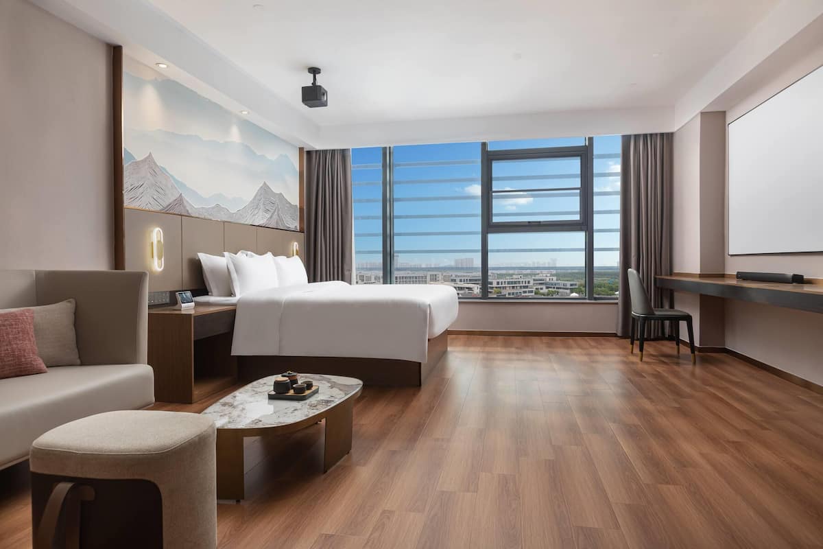 A modern hotel room with a large bed, a sofa, a desk, and a view of the city through large windows.