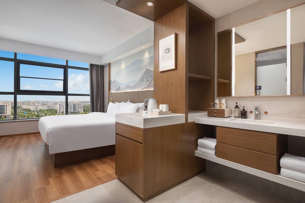 A modern hotel room with a large bed, a wooden vanity, a large mirror, and a city view.