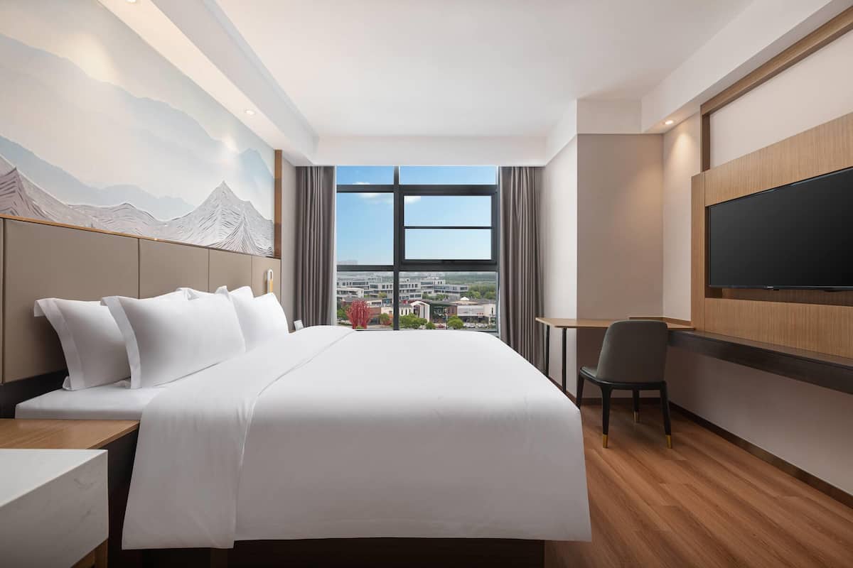 A hotel room with a large bed, a desk, a chair, a flat-screen TV, and a large window offering a city view.