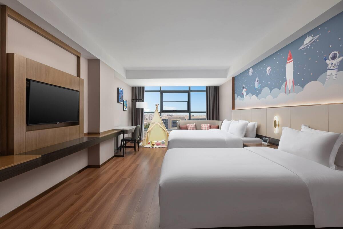 A hotel room with a large flat-screen TV, a desk with a chair, two beds, a small tent, and a mural of space-themed artwork on the wall.