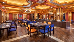 Amplia zona de comedor de hotel con largas mesas y sillas azules, un bar central y detalles ornamentales en el techo.