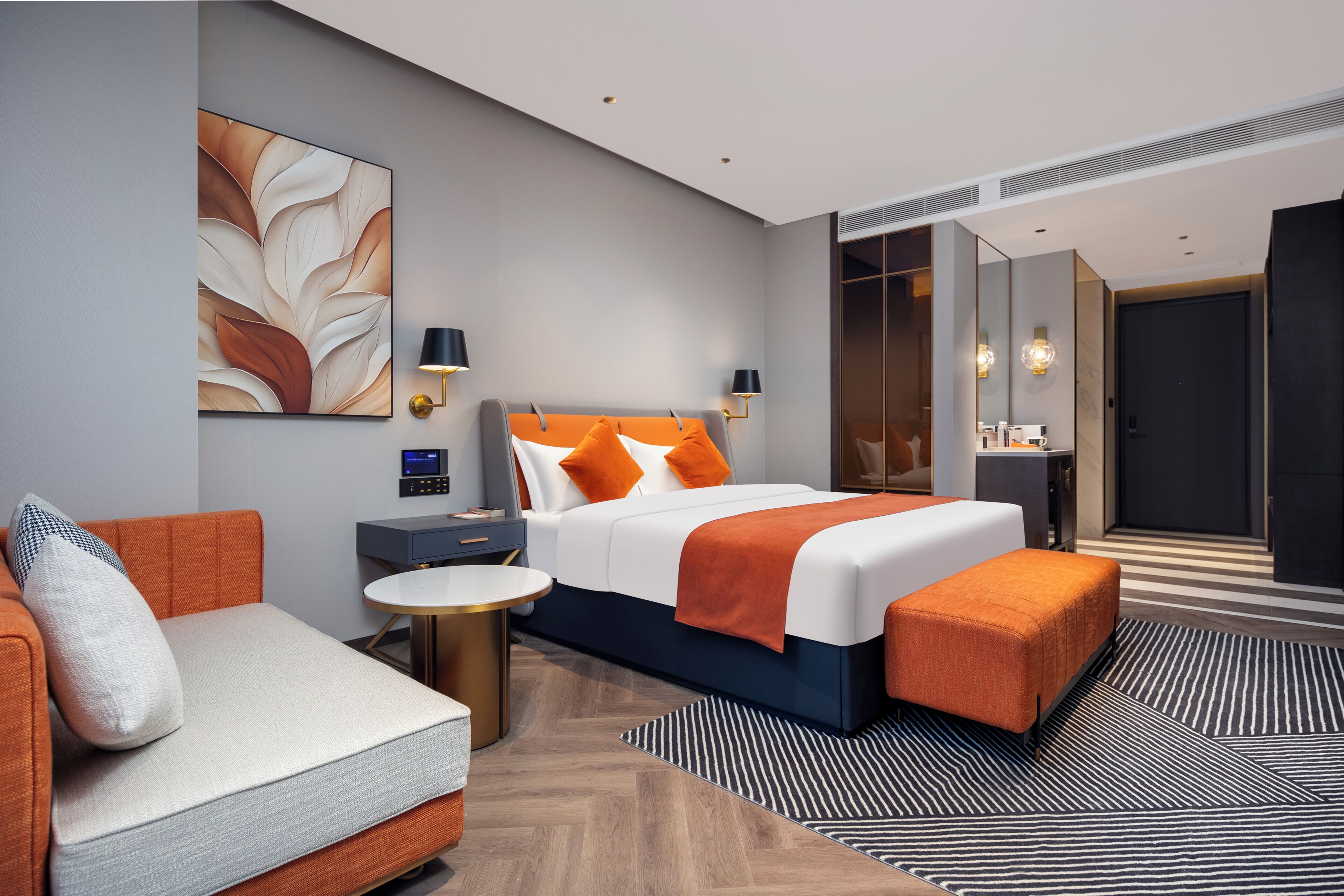 A modern hotel room with a bed, a sofa, a bedside table, a lamp, a digital clock, and a large mirror.