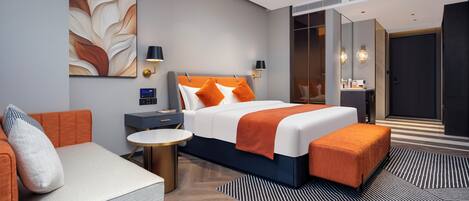 A modern hotel room with a bed, a sofa, a bedside table, a lamp, a digital clock, and a large mirror.
