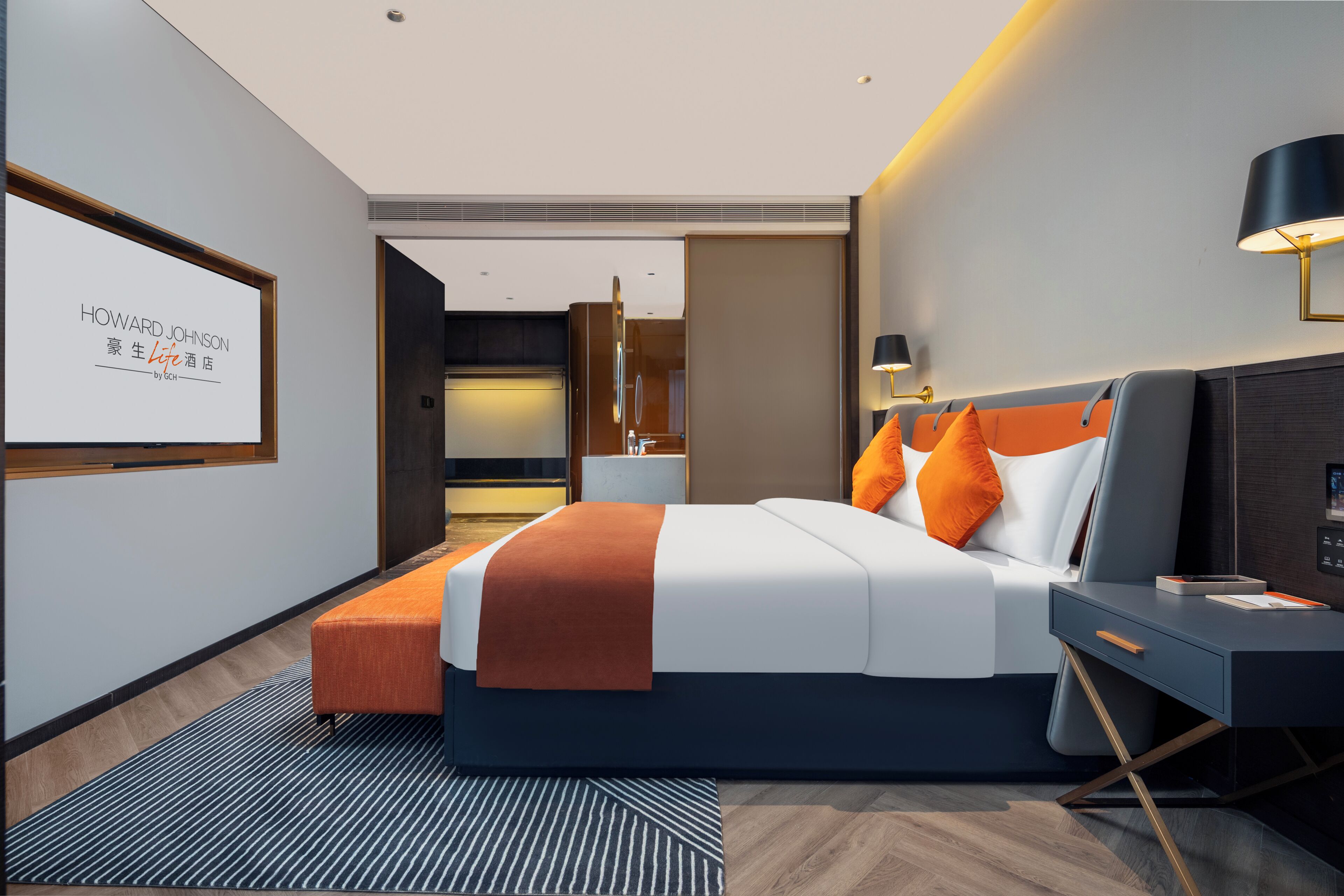 Una habitación de hotel moderna con una cama grande, un escritorio y un televisor mostrando 'HOWARD JOHNSON'.