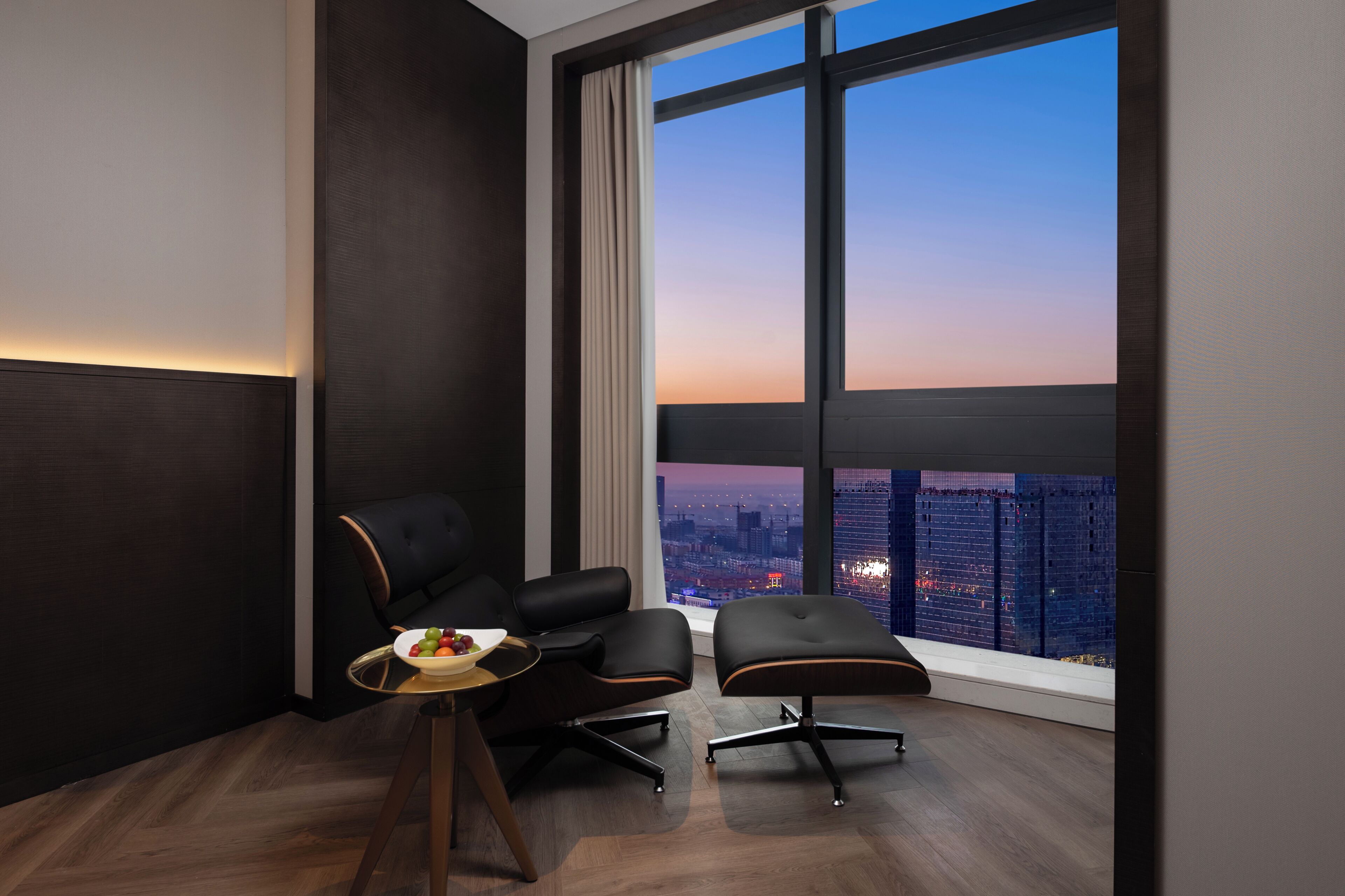 A modern room with a large window, a desk, two chairs, and a small table with a bowl of fruit.
