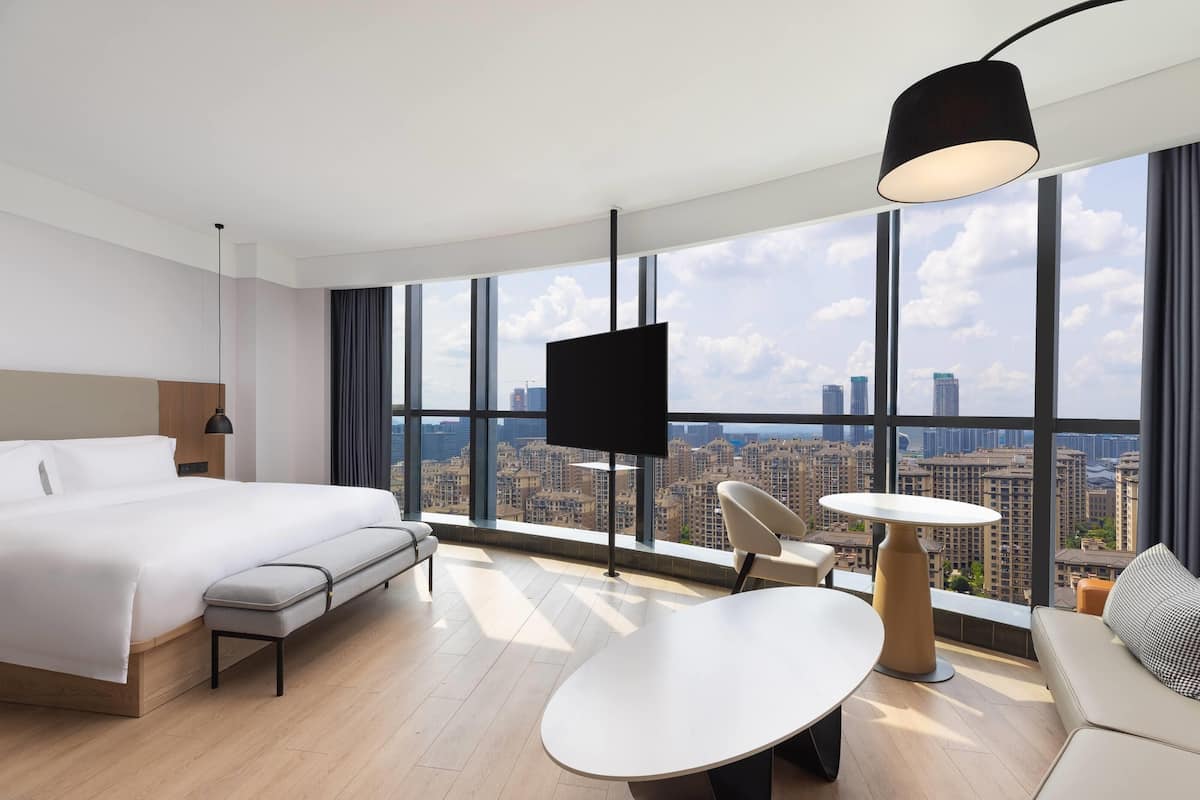Habitación de hotel moderna con una cama grande, un escritorio, una silla, una mesa redonda y un televisor. La habitación tiene ventanas amplias con vista a la ciudad.