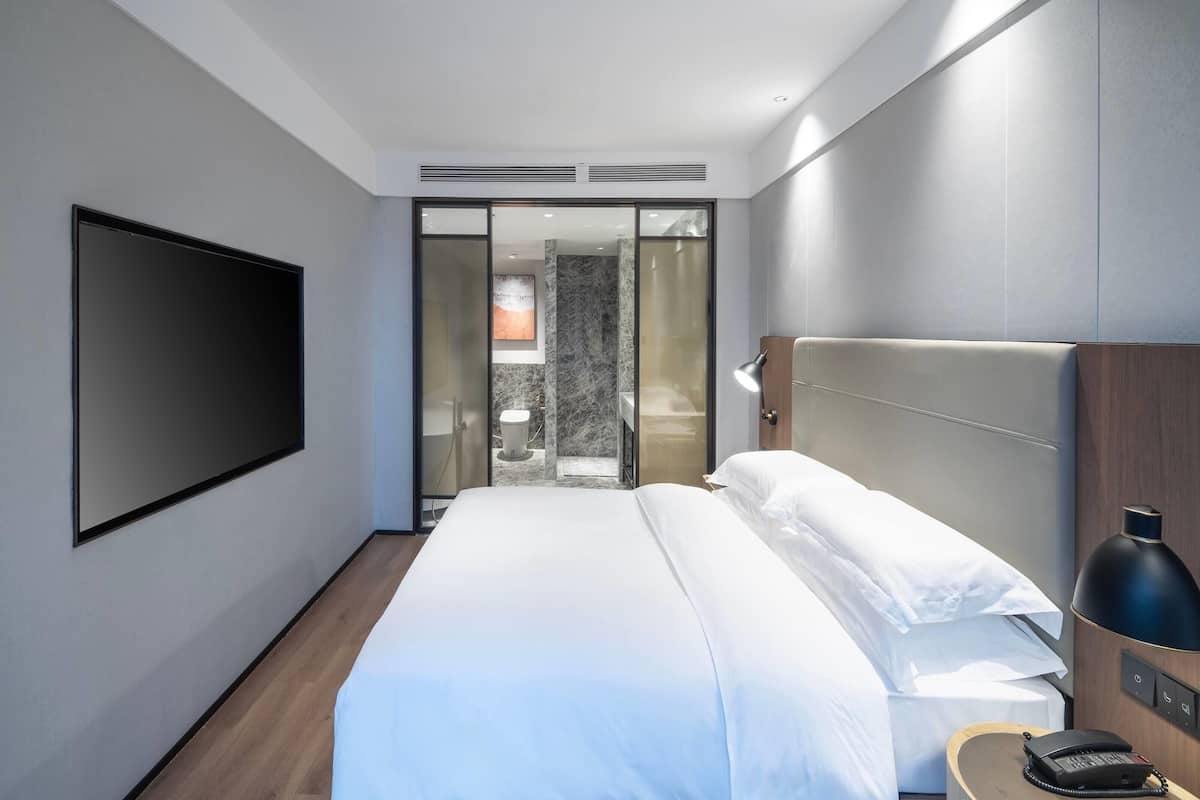 Una habitación de hotel moderna con una cama grande, un televisor de pantalla plana montado en la pared, una lámpara de noche y una mesita de noche de madera.
